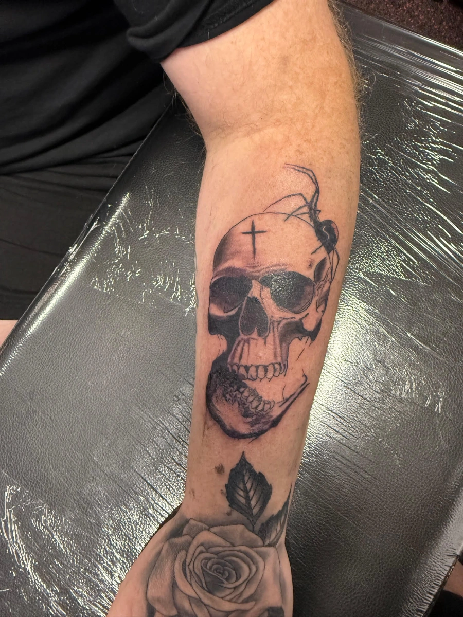 Detailed black-and-grey skull tattoo on forearm with rose and spider.