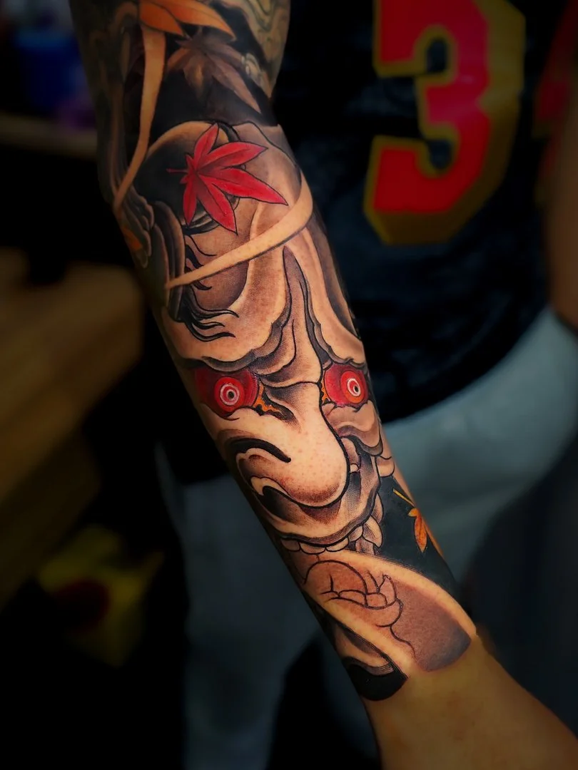 Japanese tattoo of a demon mask with red eyes and leaves.