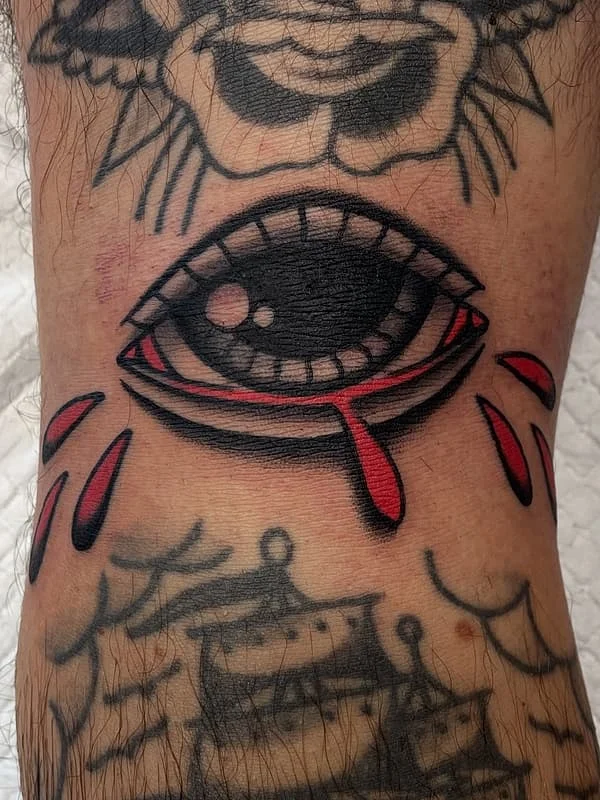 Traditional eye tattoo with red tears on leg.