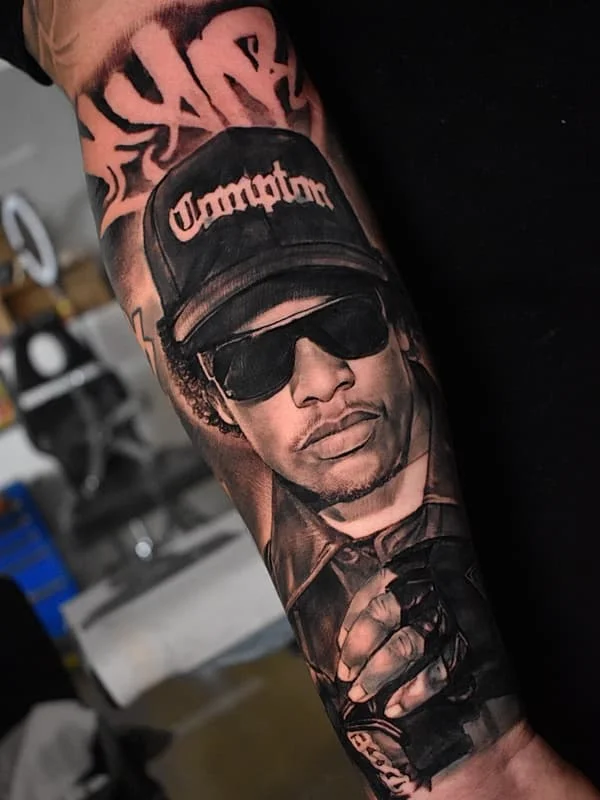 Black-and-grey portrait tattoo of a hip-hop artist on forearm.