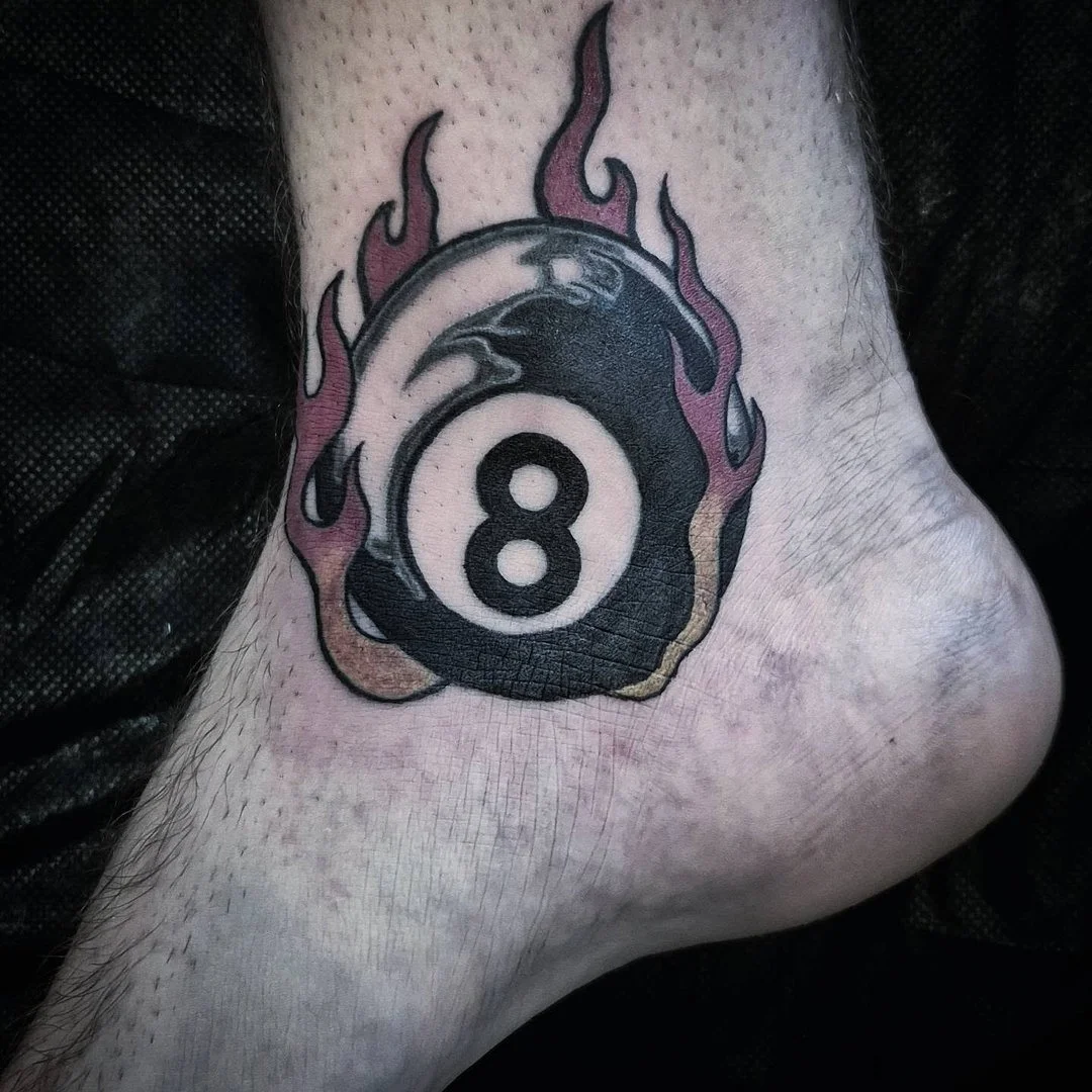 Small traditional eight-ball tattoo with flames on ankle placement
