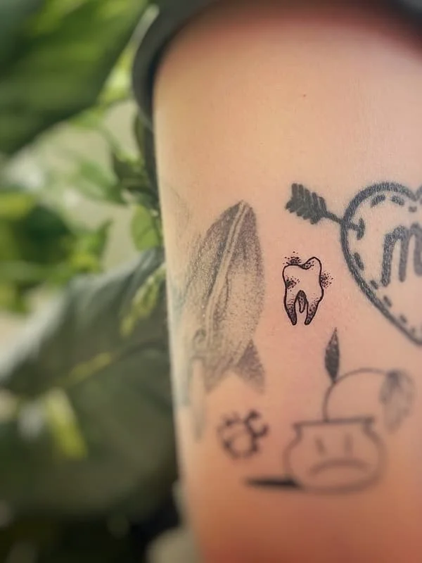 Minimalist black-grey tattoo on arm featuring a tooth and heart.