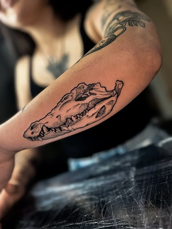 Illustrative blackwork tattoo of a crocodile skull on forearm.