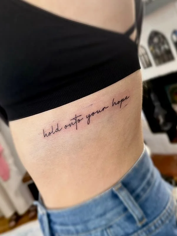 Script tattoo on rib saying 'hold onto your hope'.
