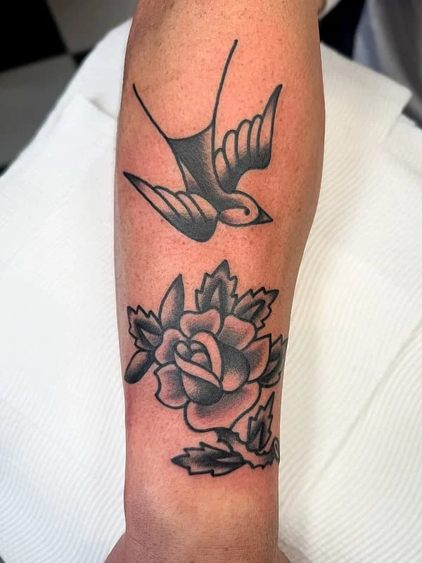 Traditional black-and-grey tattoo of a bird and rose on forearm.