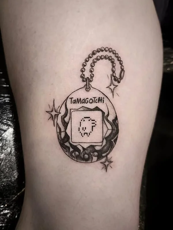 Illustrative Tamagotchi tattoo on arm in black-grey ink.