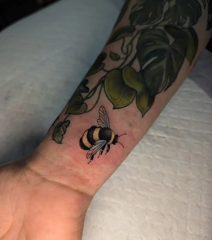 Colorful bee tattoo on forearm with green leaves.
