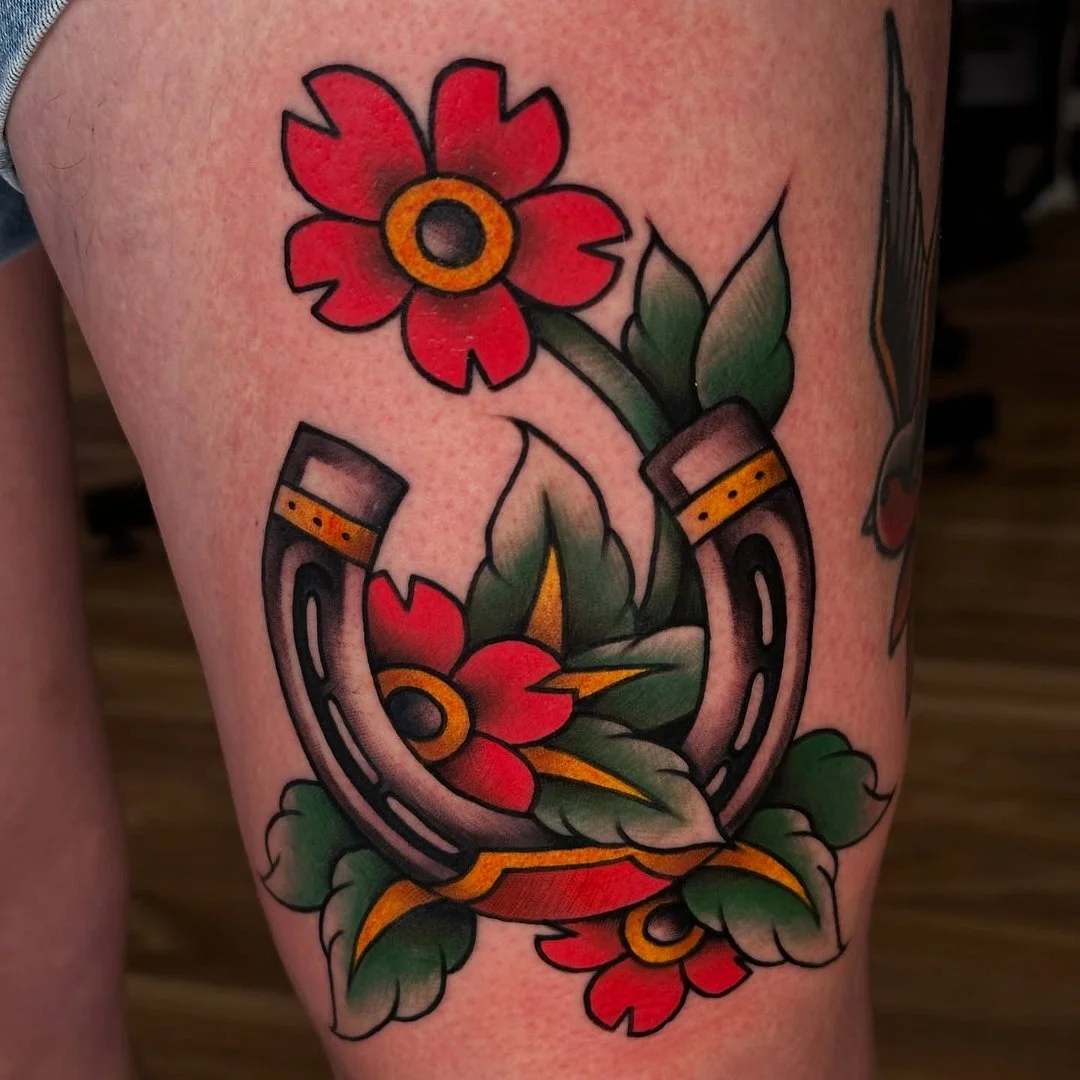 Colorful traditional tattoo of a horseshoe and flowers on thigh.