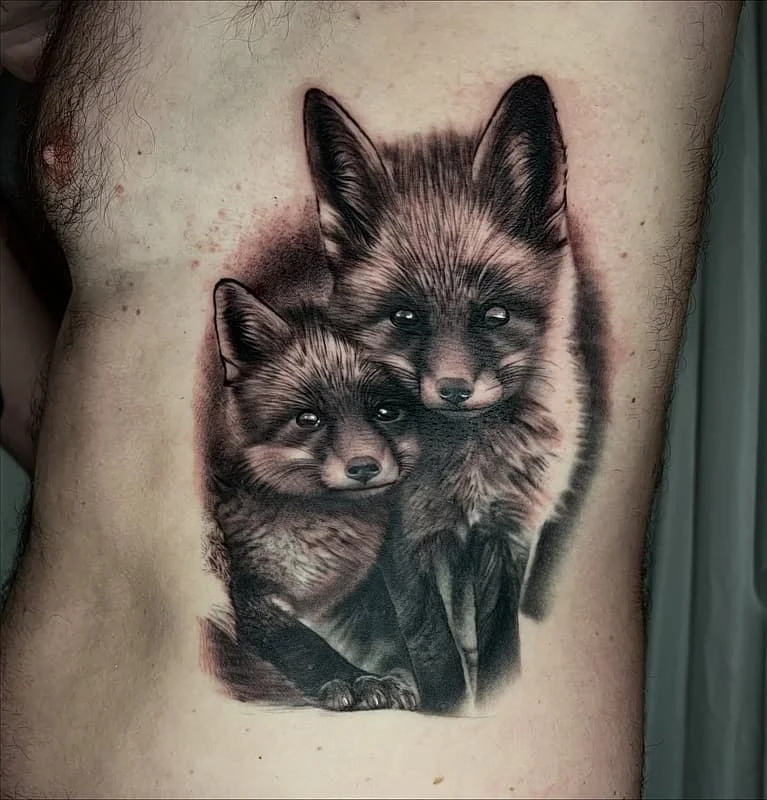 Realistic black and grey tattoo of two foxes on rib.