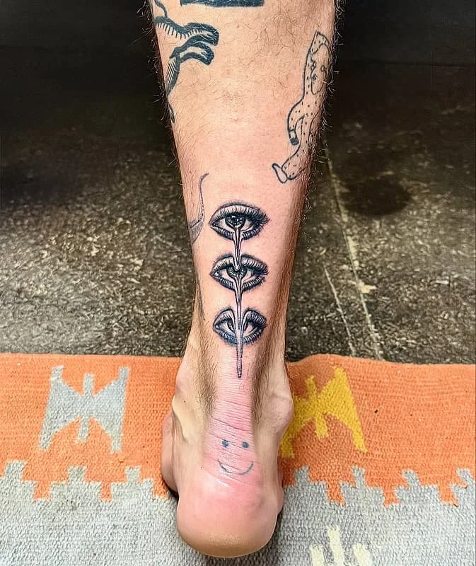 Black-and-grey illustrative tattoo of eyes and flowers on ankle.