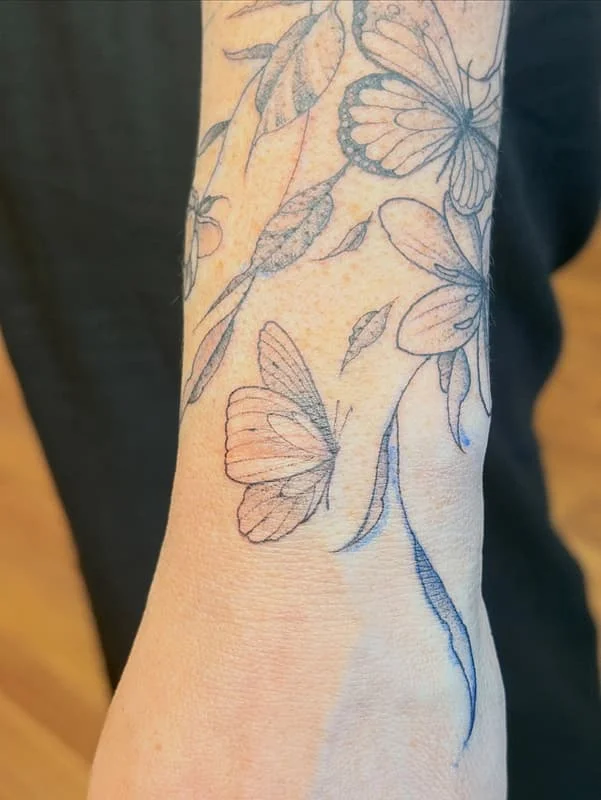 Fine-line tattoo of butterflies and flowers on forearm.