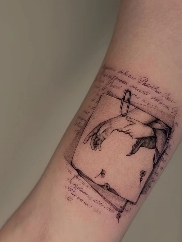 Illustrative black and grey forearm tattoo of hands and paper