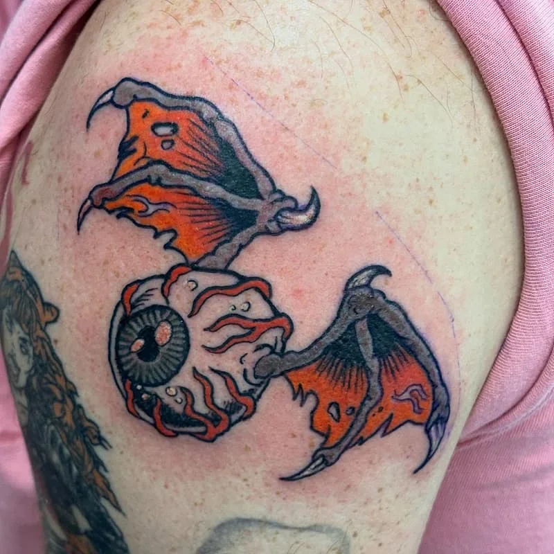Colorful illustrative tattoo of an eye with wings on shoulder.